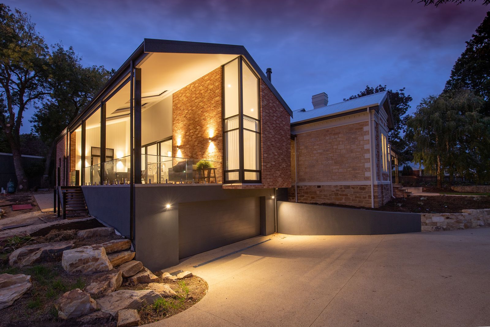 Modern glass-and-timber house exterior at dusk with warm interior lighting glowing through the windows
